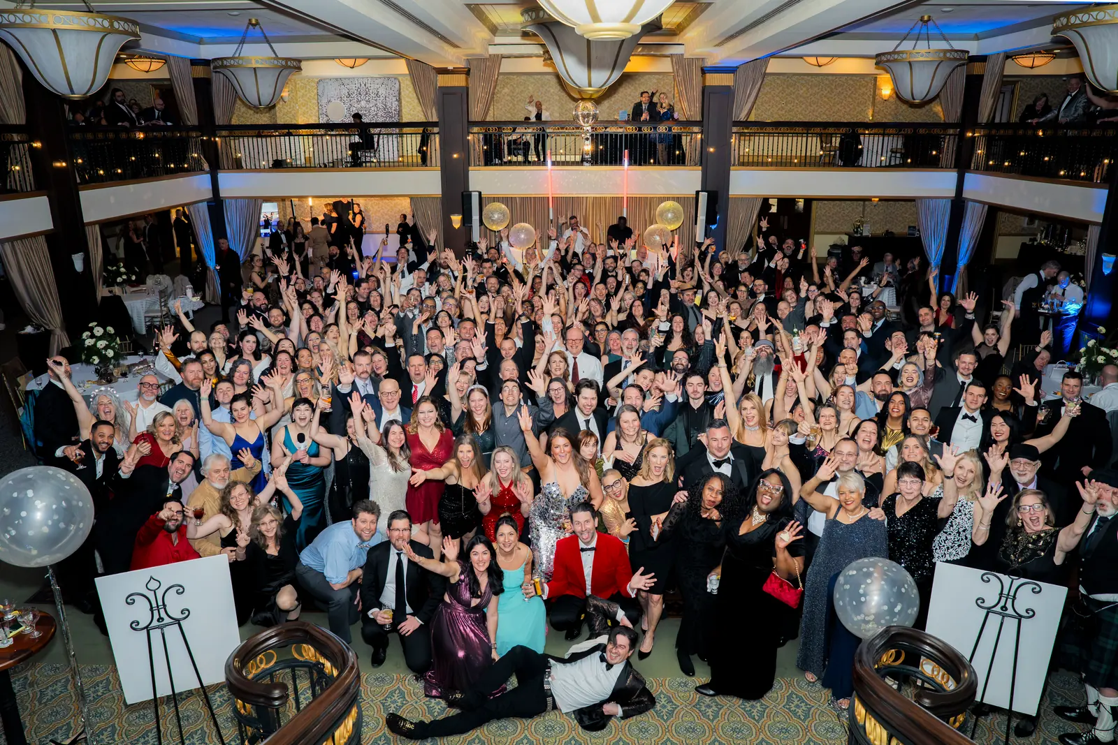 A packed two-level ballroom of gala guests in evening wear waving up at the camera from the balcony
