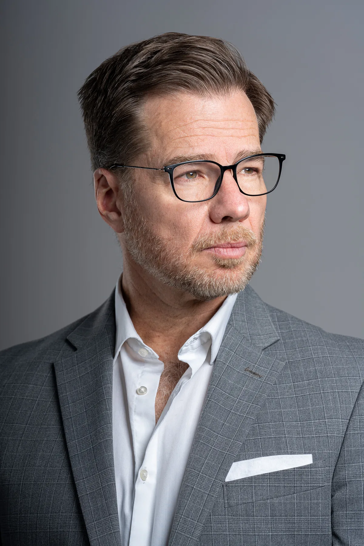 Studio portrait of a man in glasses and a grey check suit jacket against a grey backdrop, looking off camera