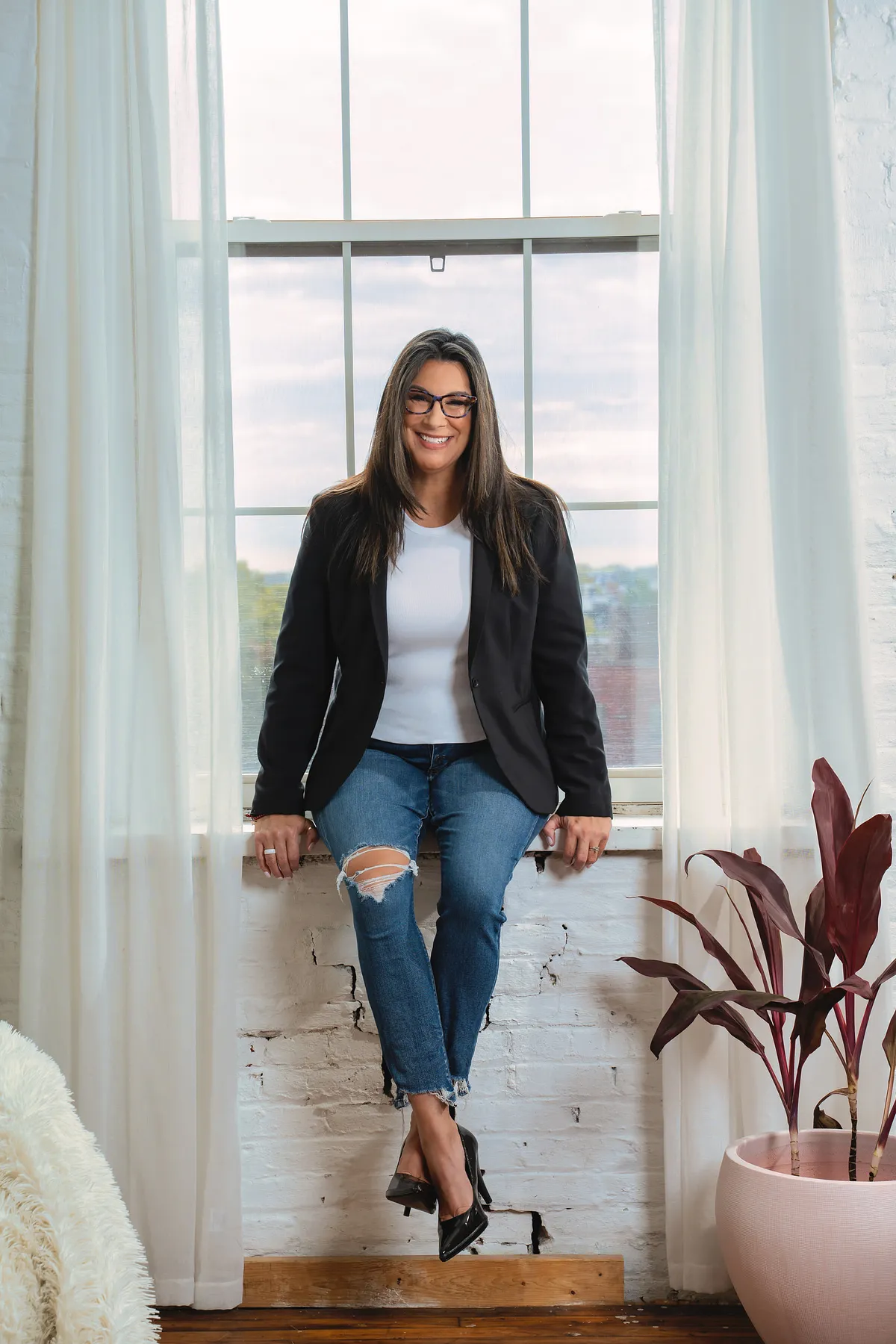 Professional in a black blazer and jeans seated on a loft window ledge between white curtains